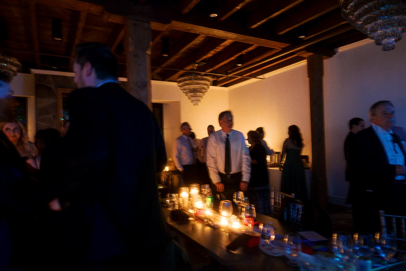 Guests mingling indoors around candlelit tables beneath chandeliers.