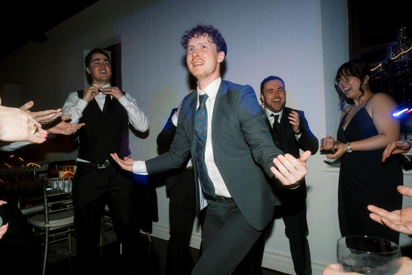 Man in a suit dancing while guests clap around him at an indoor celebration