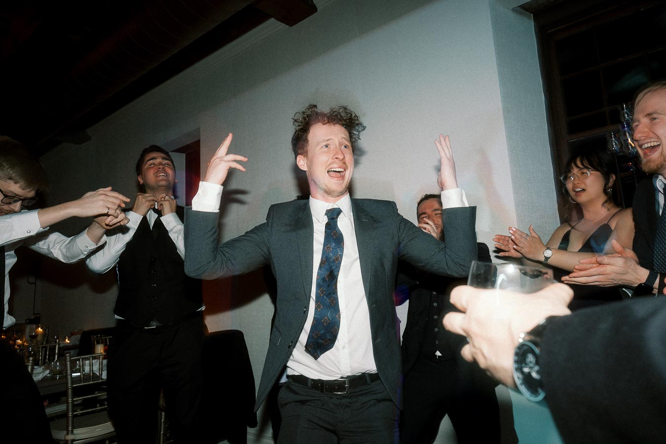 Man in a suit dancing while friends clap around him at an indoor celebration