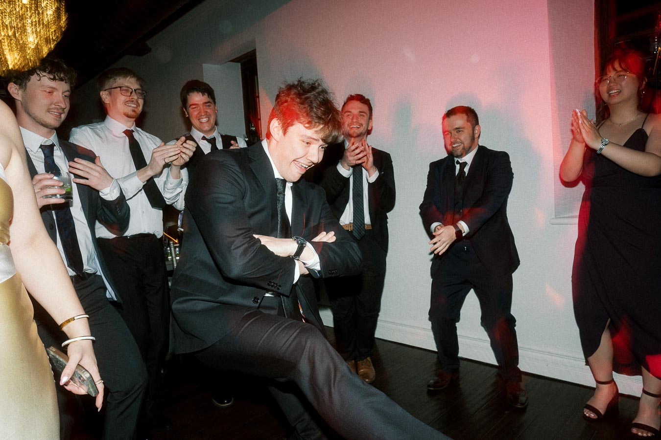 Man in a black suit dancing as guests clap around him indoors