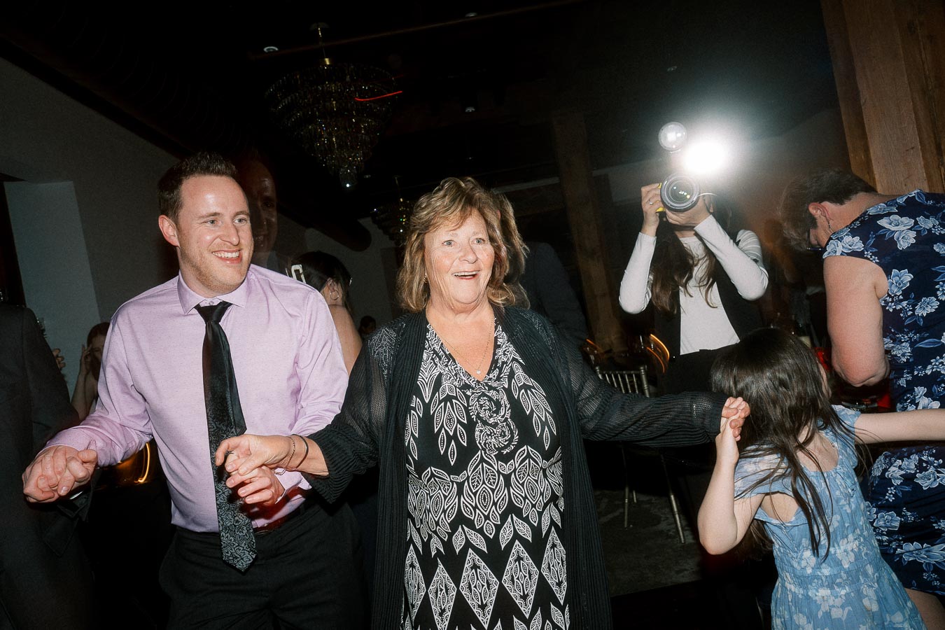 Guests holding hands and dancing indoors while a photographer uses flash nearby