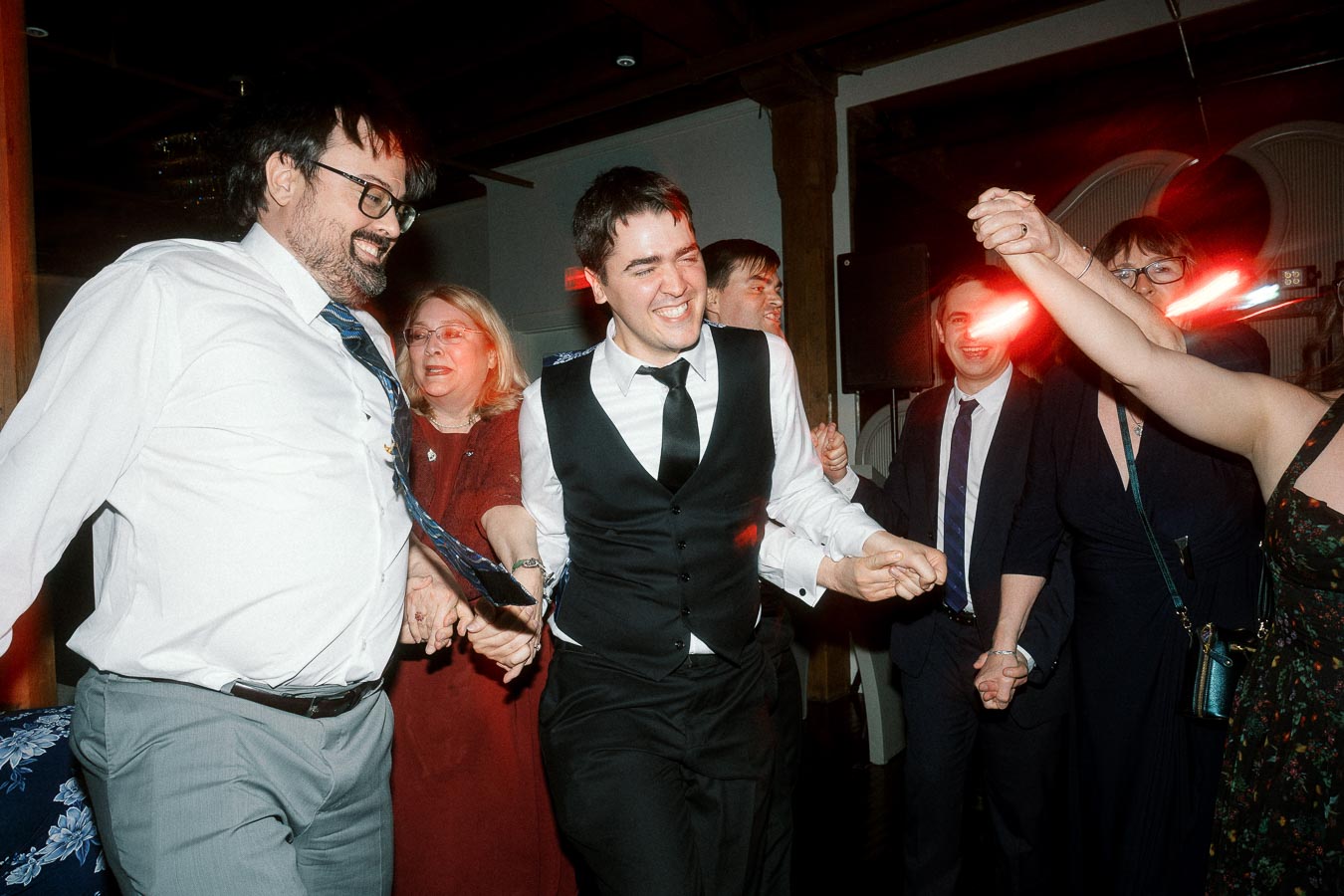 Group of formally dressed guests holding hands and dancing indoors with red party lights