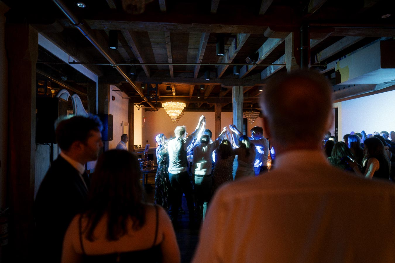 Guests dancing indoors beneath wooden beams and chandeliers at a lively celebration.
