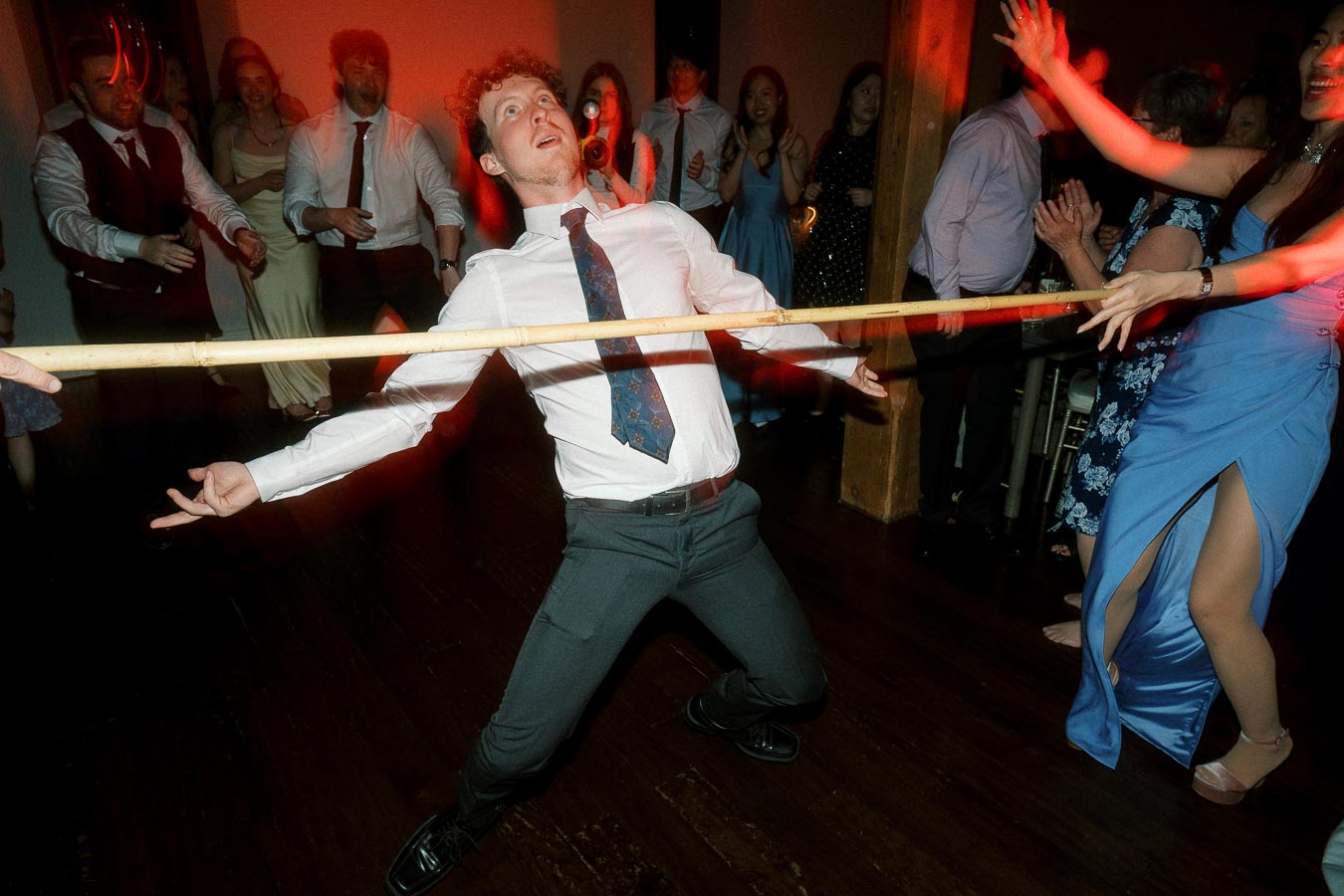 Man in dress shirt and tie limbo dancing under a pole at a lively reception