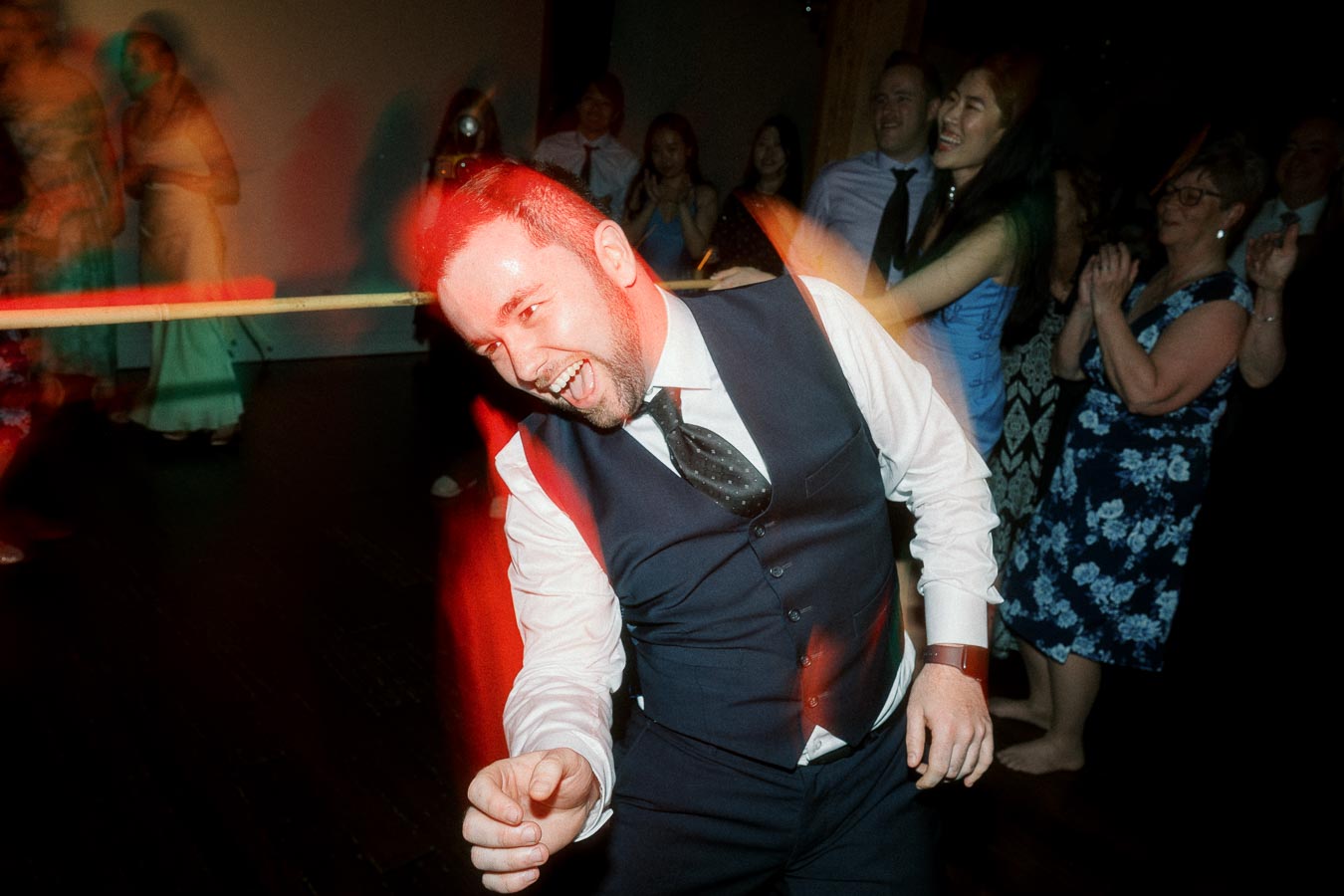 Man in a vest laughing and dancing among guests on a dark dance floor