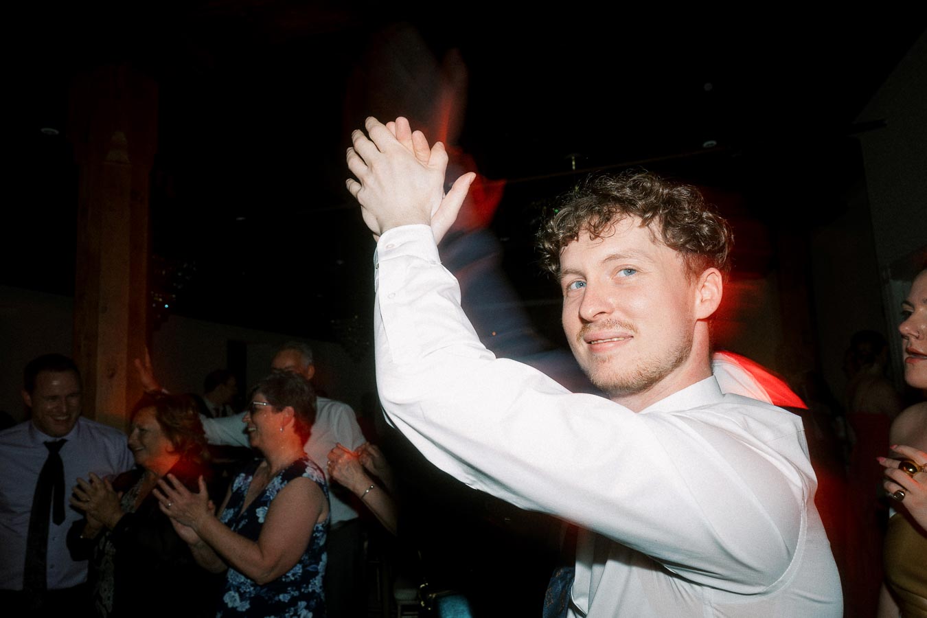 Man in a white shirt clapping on a dark dance floor with guests nearby