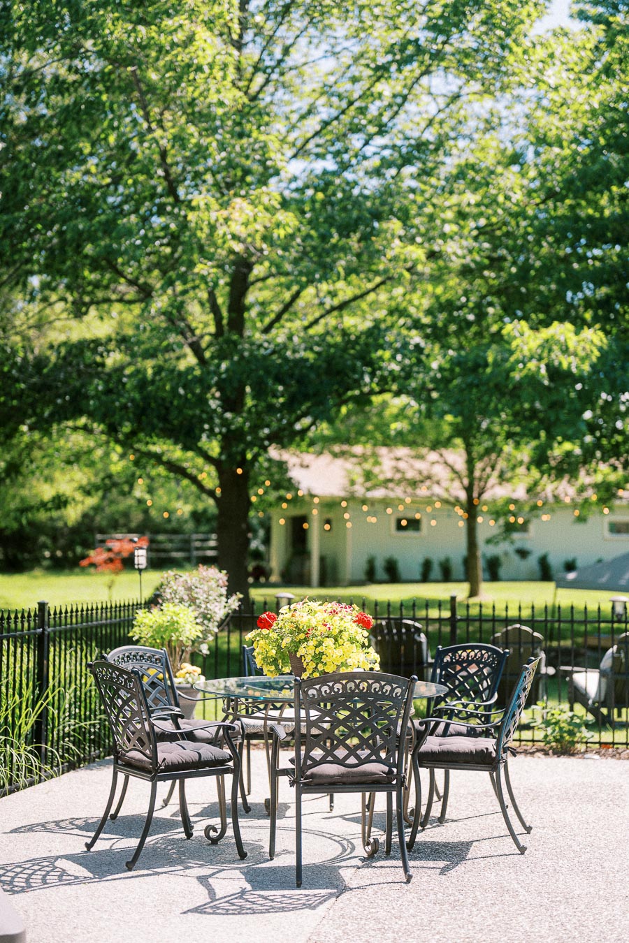Outdoor patio with black wrought iron furniture and a round glass table, featuring a vibrant flower arrangement. Surrounded by lush greenery and a fenced garden, with string lights enhancing the peaceful and inviting atmosphere. Perfect setting for outdoor relaxation or dining.
