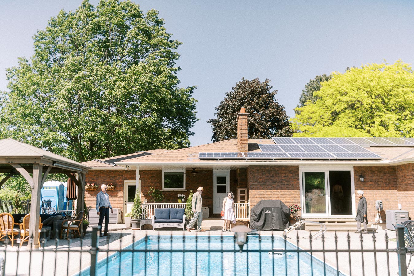 Modern brick house with solar panels on the roof, a backyard oasis featuring a swimming pool, gazebo with outdoor seating, and lush green trees for a relaxing atmosphere.