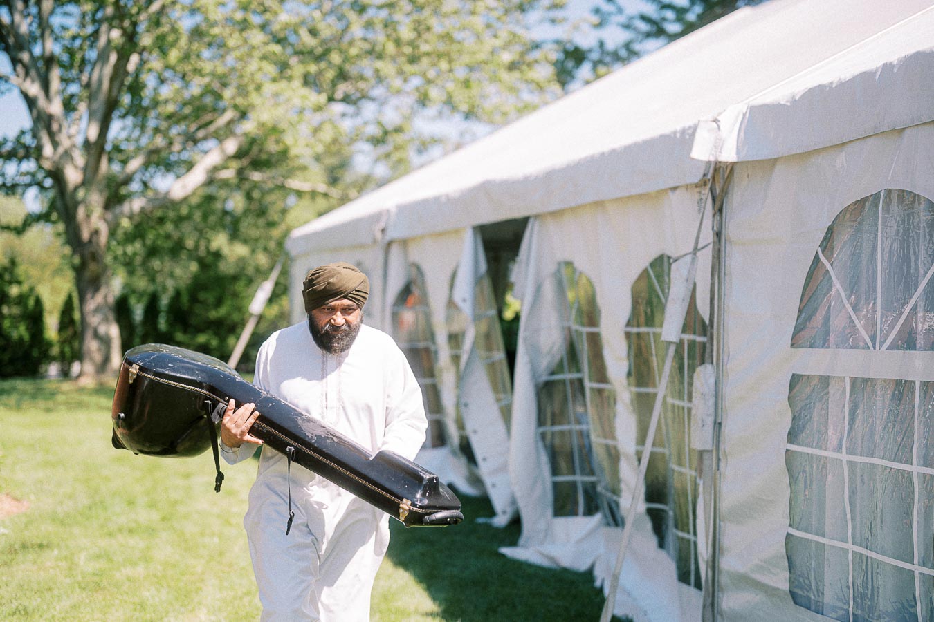 Man in traditional attire carrying a large instrument case near a white event tent surrounded by greenery on a sunny day.