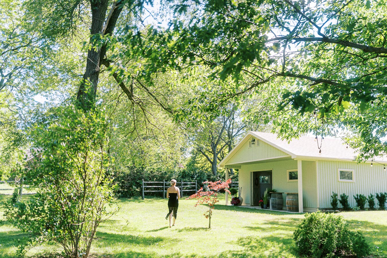 A woman walks through a sunlit garden towards a small, charming shed surrounded by lush green trees and plants, under a clear blue sky.