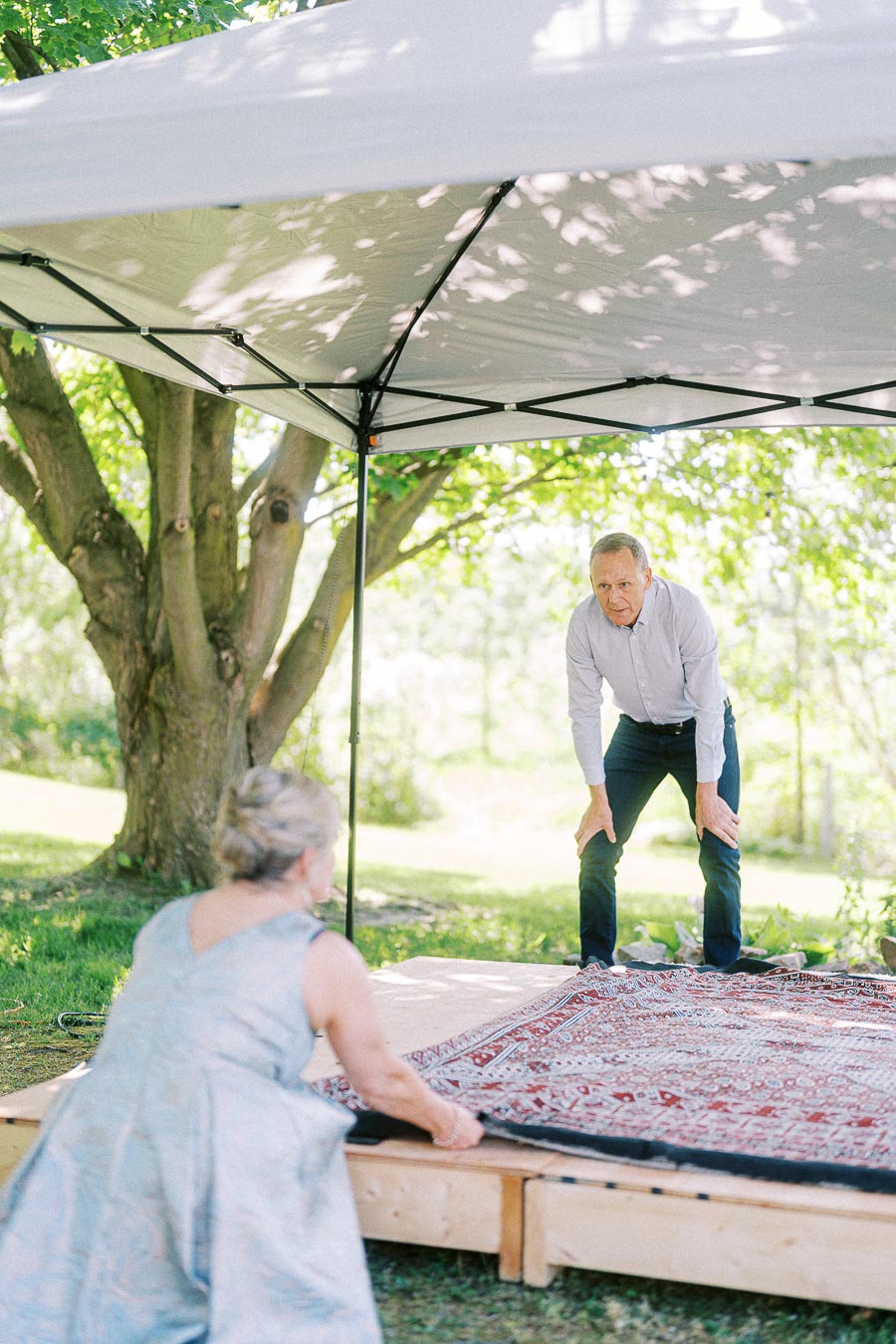 Elderly couple setting up an outdoor canopy and arranging a decorative rug, under the shade of a large tree in a sunny garden setting.