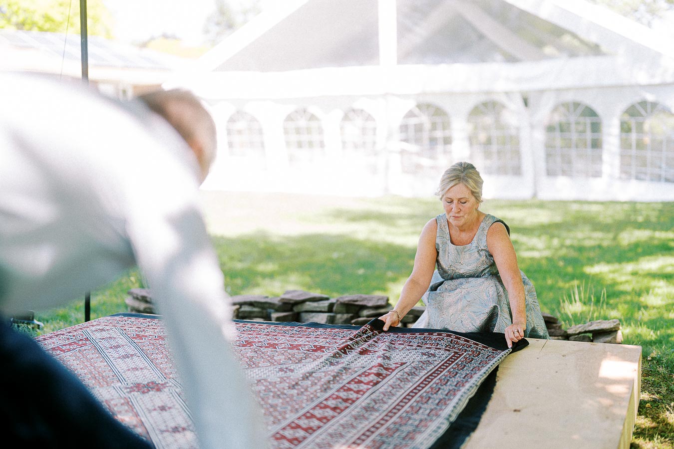 A woman carefully adjusts a patterned rug on a wooden platform outdoors, with a white event tent in the background on a sunny day.