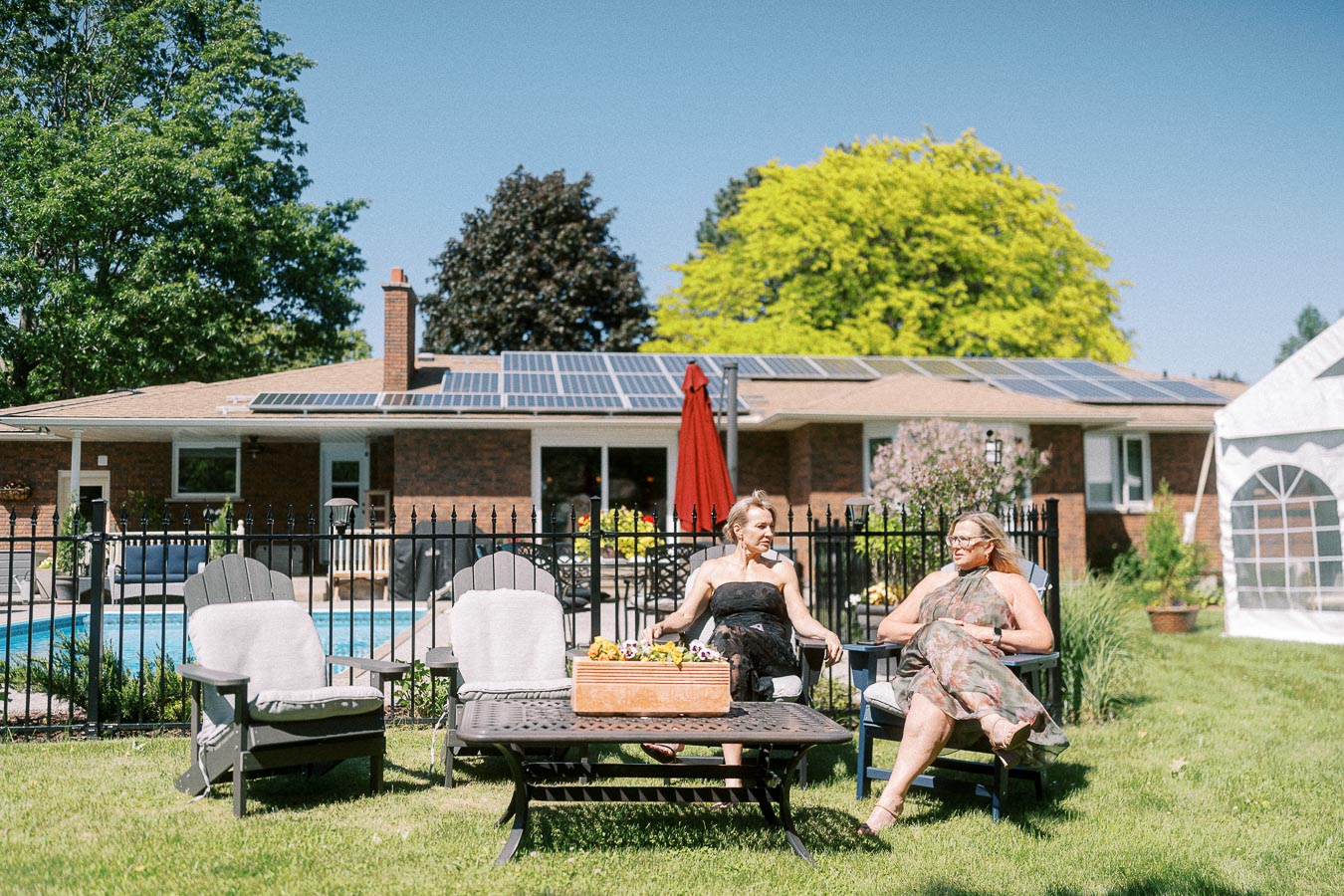 Two women relaxing in a sunny backyard with solar panels on the house roof in the background. The outdoor setting includes a pool, patio furniture, and lush greenery, illustrating a sustainable and leisurely lifestyle.
