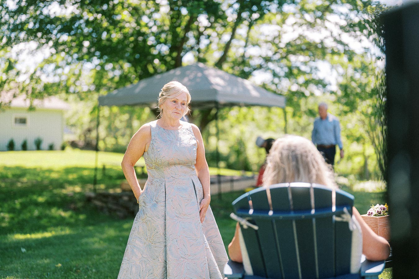 Elegant woman in a silver dress standing outdoors at a garden event, interacting with a seated person in a blue chair. Sunlit trees and a canopy create a serene background.
