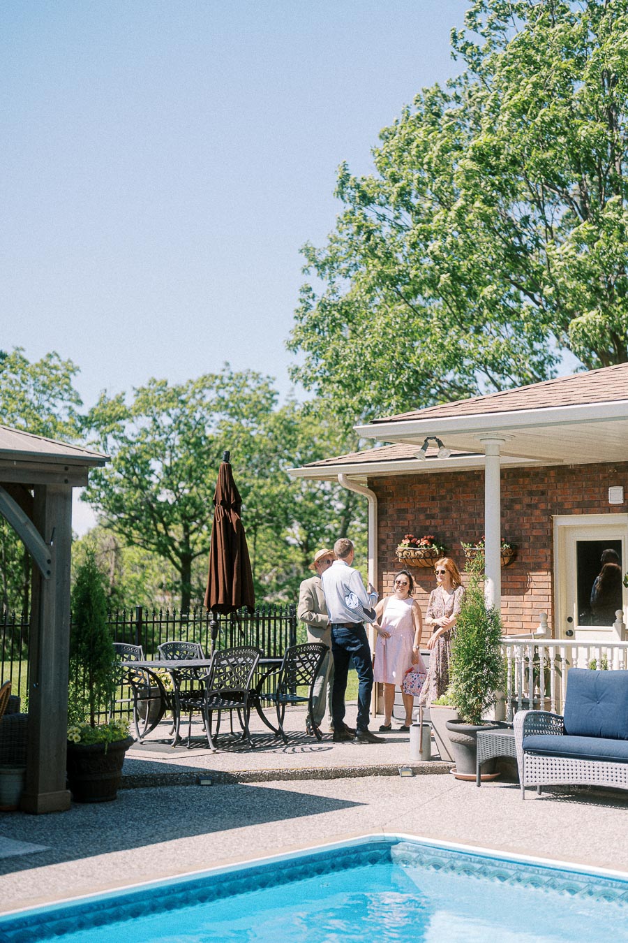 Outdoor gathering by a poolside patio with a group of people socializing under clear blue skies, surrounded by lush green trees and cozy furniture.
