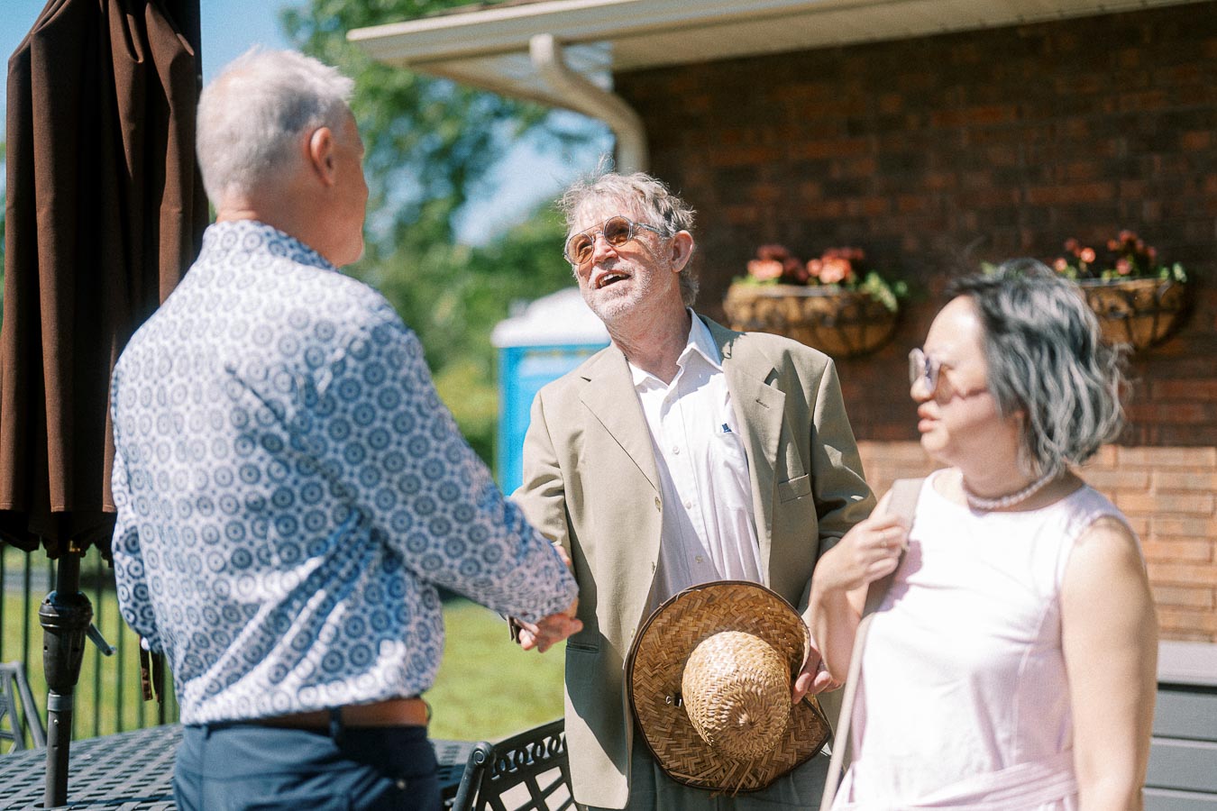 Elderly man in sunglasses shakes hands with another man in a patterned shirt while a woman in sunglasses stands beside them, outdoors with a building and flowers in the background on a sunny day.