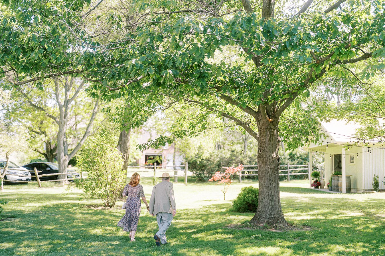 Elderly couple walking hand in hand through a lush green garden under a large tree, with a quaint cottage and string lights in the background, evoking a sense of tranquility and togetherness in a natural setting.