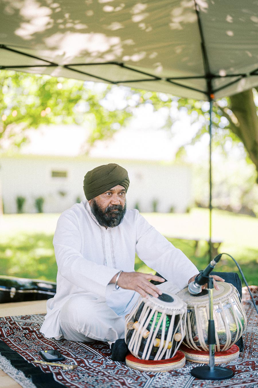 A man wearing a turban plays traditional tabla drums under a canopy, surrounded by a lush green garden, highlighting cultural music performance outdoors.