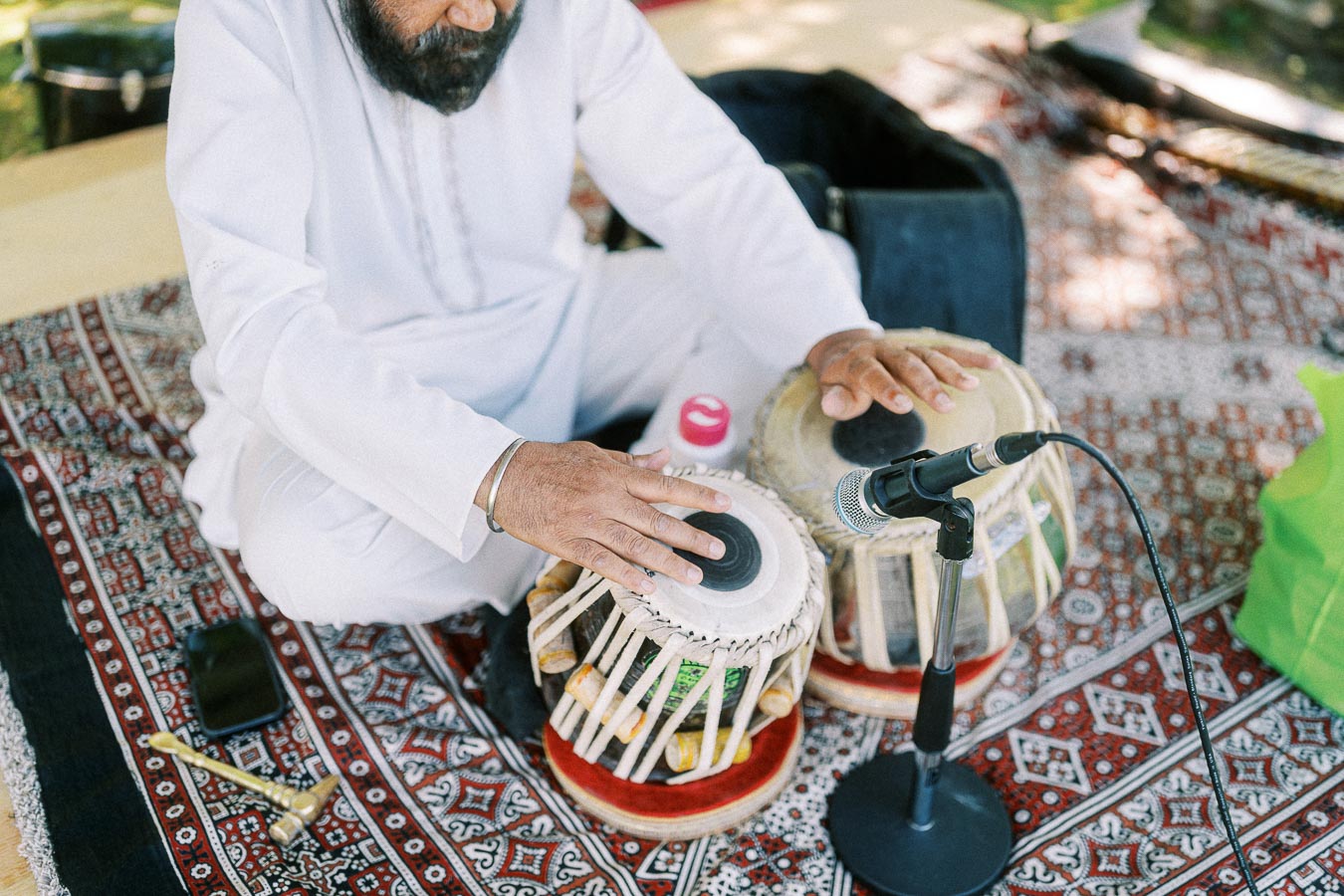 Musician playing traditional Indian tabla drums on a patterned rug with a microphone, outdoors.