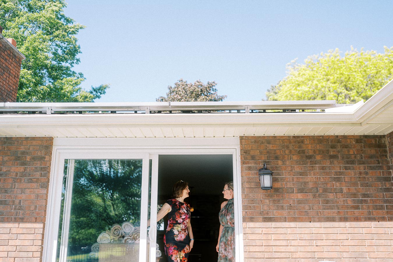 Two women standing in the entrance of a brick house, engaged in conversation. The scene is set against a backdrop of a clear blue sky and lush green trees, highlighting a serene outdoor atmosphere.