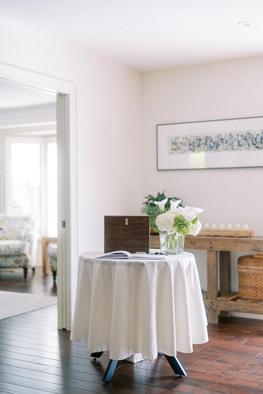 Elegant living room corner featuring a round table with a white tablecloth, adorned with a vase of white flowers and a wooden box, with a painting on the wall and an open doorway showing a cozy sitting area in the background.