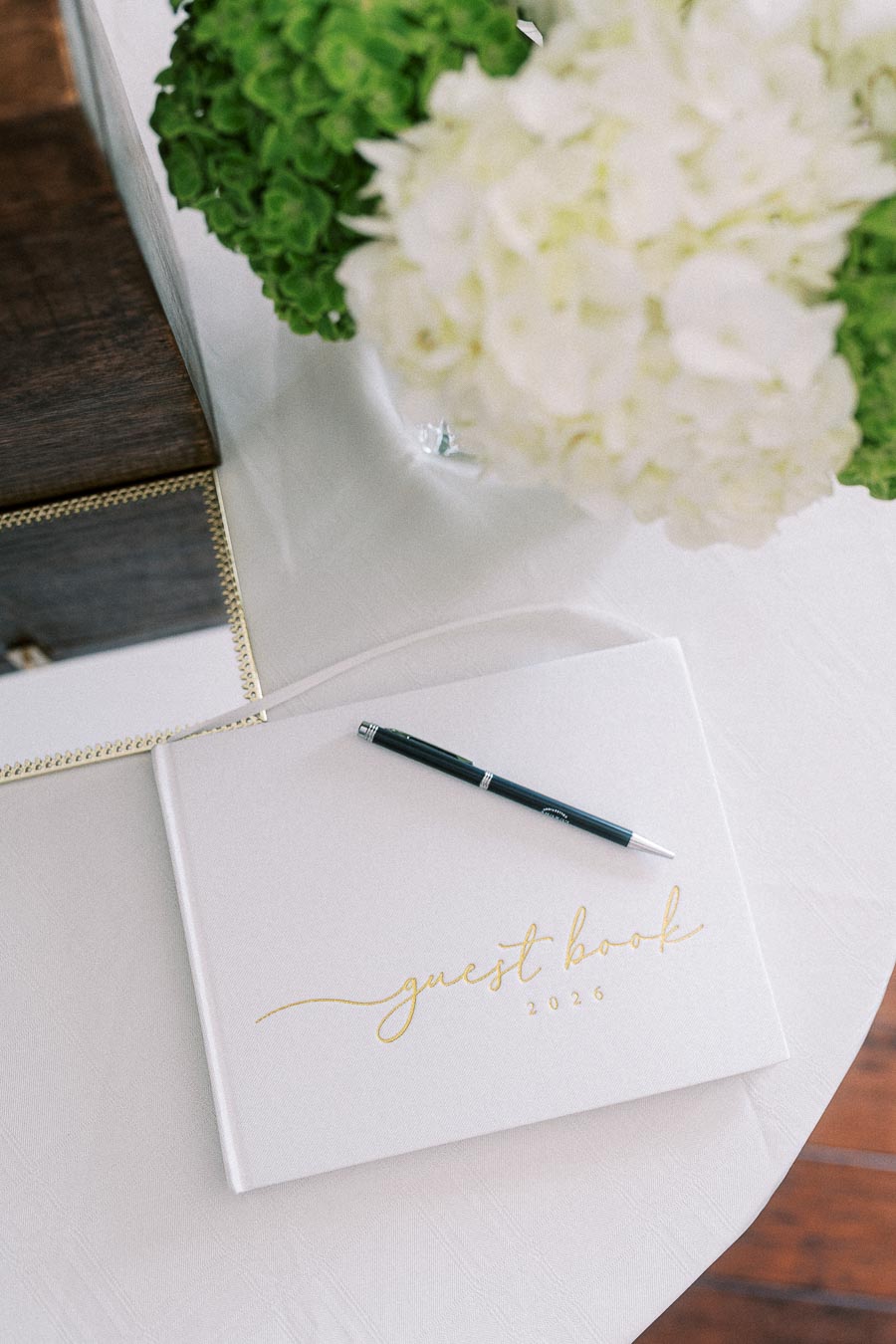 Elegant 2026 guest book with a black pen on a white table, surrounded by lush green and white flowers. Perfect for capturing memorable moments at special events.