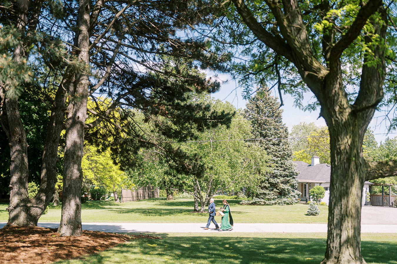 A couple walking on a path through a lush park on a sunny day, surrounded by tall trees and greenery, with a house visible in the background.