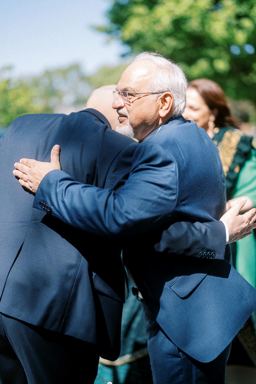 Two men in formal blue suits embrace warmly outdoors during a sunny day, symbolizing friendship and connection at a social gathering.