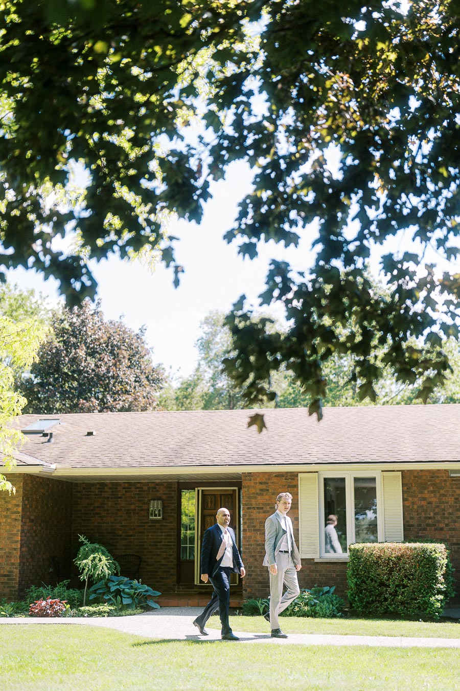 Two people walk along a path in front of a brick house on a sunny day, surrounded by lush greenery and trees.