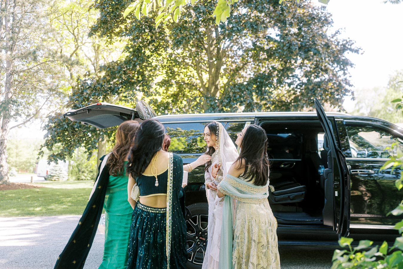 Wedding celebration with a bride surrounded by friends in elegant traditional attire, preparing to enter a black car under a sunny, lush green setting.