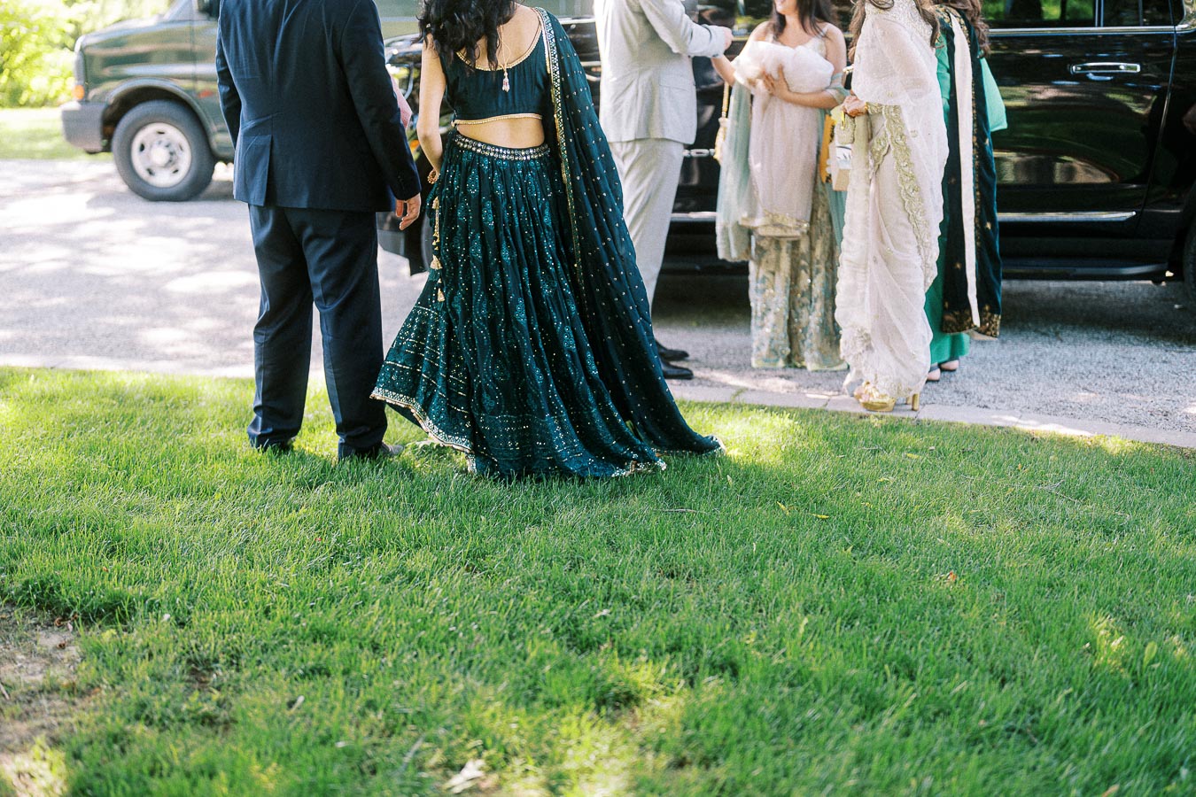 A group of people dressed in formal and traditional South Asian attire stand on a grassy area next to a black vehicle, possibly attending an outdoor event or celebration.