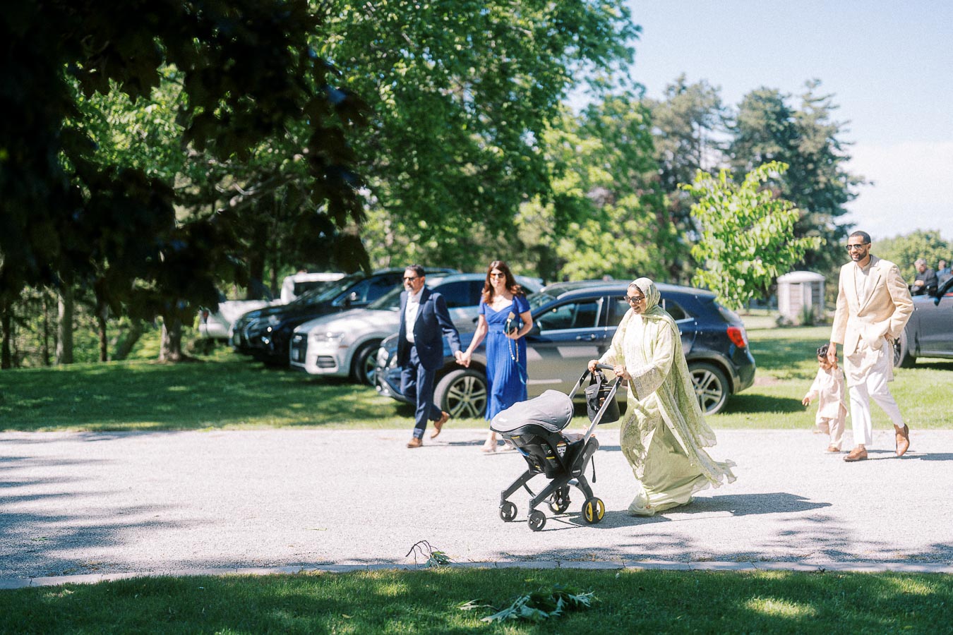 A group of people leisurely walking on a sunny day in a park, with a woman in a green dress pushing a stroller and others dressed elegantly nearby, cars parked in the background under lush green trees.
