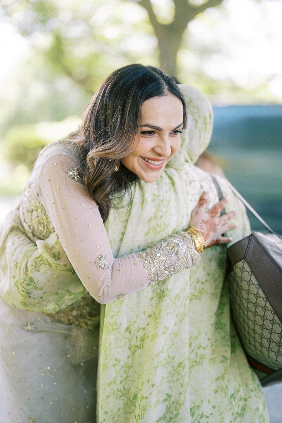 Smiling woman in elegant traditional attire hugs another person outdoors, showcasing joy at a cultural event.