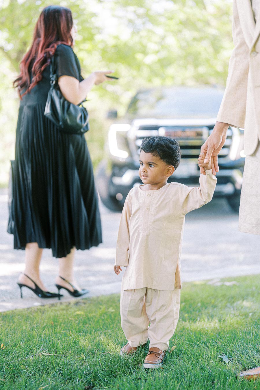 A young boy in traditional attire holding an adult's hand, standing on a grassy area with people and a parked car in the background.