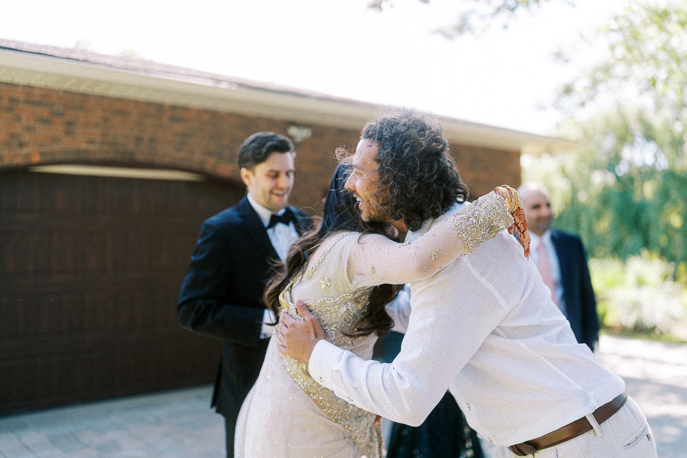 Outdoor celebration with people hugging in formal attire, featuring festive clothing and joyful expressions, set against a sunny background with greenery and a brick building.