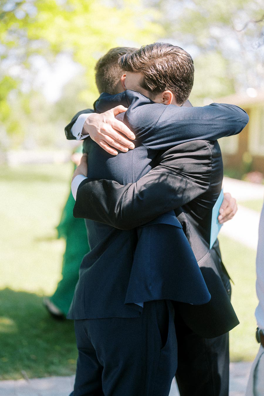 Two people in formal suits embracing warmly outdoors, surrounded by lush greenery, symbolizing friendship and celebration.