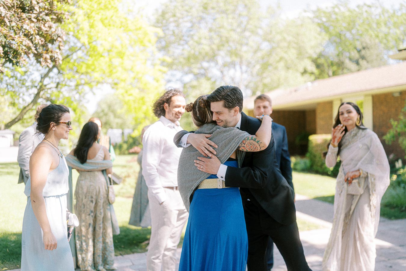 Group of people celebrating outdoors at a joyful gathering, with one person in a suit embracing another in a blue dress, surrounded by sunlight and greenery.