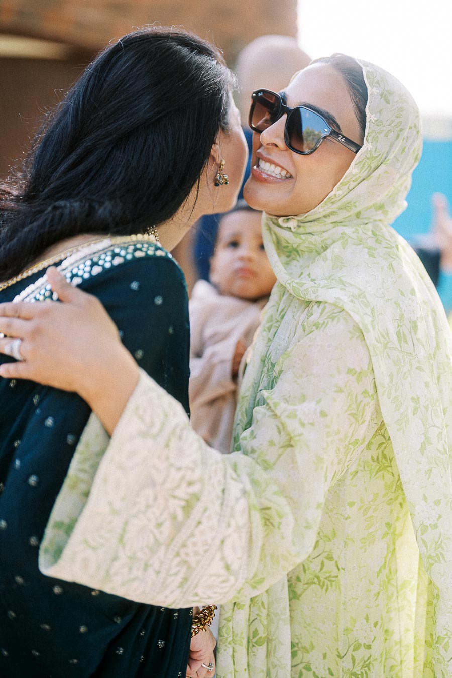 Two women in traditional attire share a warm embrace, showcasing cultural connection and happiness at a social gathering. One woman is wearing a green patterned headscarf and sunglasses, while another is in a dark, decorated dress. A baby is visible in the background, adding a family-friendly element to the scene.