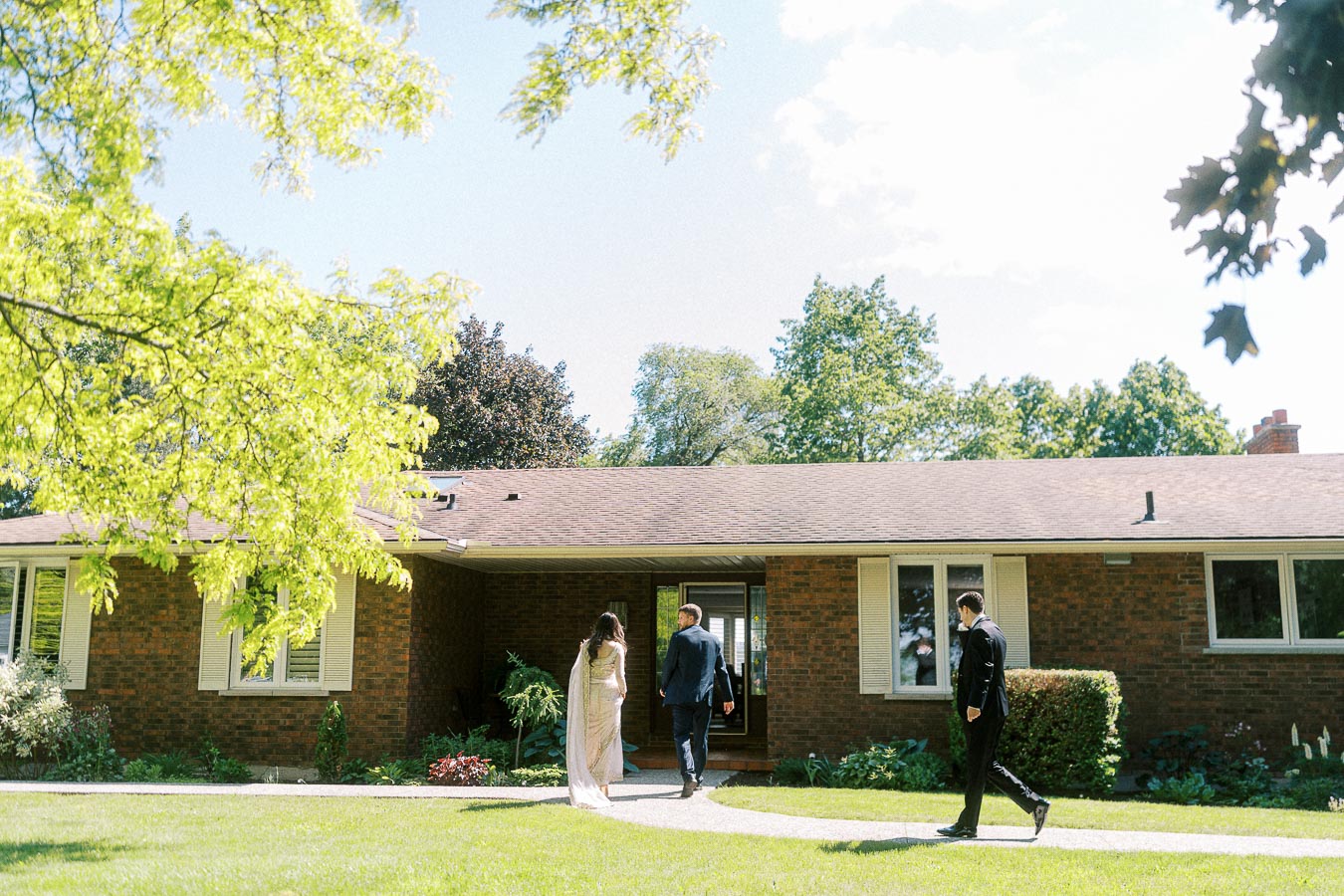 Three people walk on a path towards a cozy brick house surrounded by lush greenery on a sunny day.