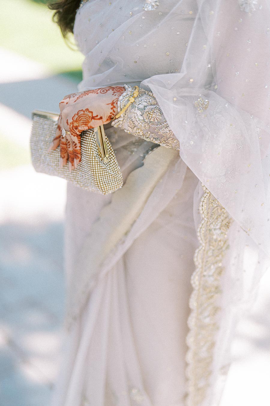 Close-up of a person wearing an elegant white embroidered saree with intricate detailing, holding a beaded clutch. The person has henna adorned hands, adding a traditional touch to the sophisticated attire.