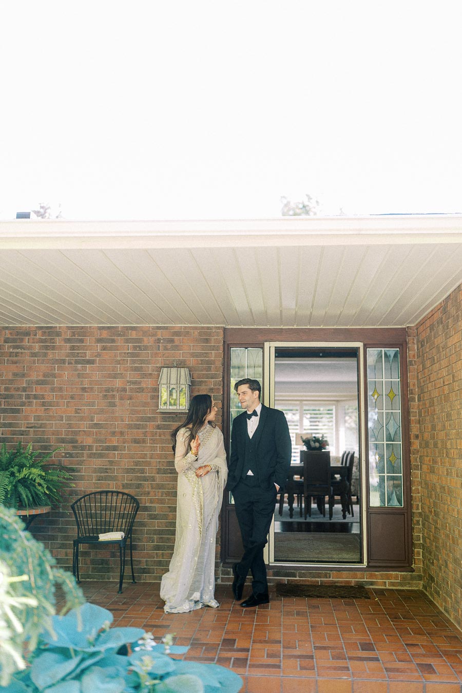 Elegant couple standing on patio in front of brick house, both dressed in formal attire, smiling at each other as the sunlight filters through the trees.