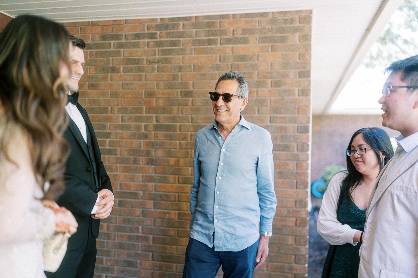 Group of people having a conversation outside a brick building. One person is wearing sunglasses and a casual outfit, while others are dressed in formal attire, including a man in a suit. They appear to be enjoying a social gathering on a sunny day.
