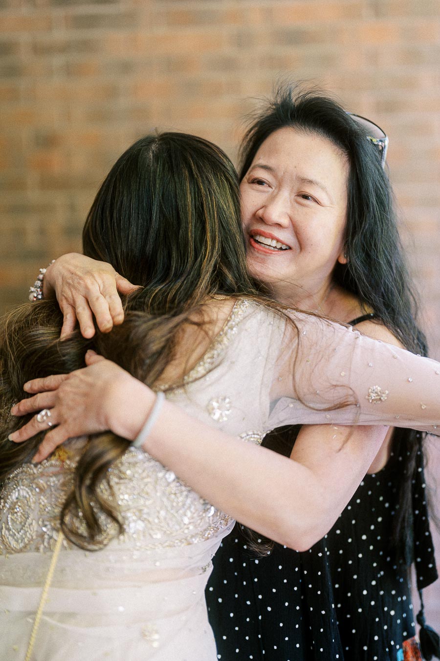 Two women embracing warmly, one in a sequined dress and the other in a black polka dot outfit, captured against a brick wall background, highlighting joy and connection.