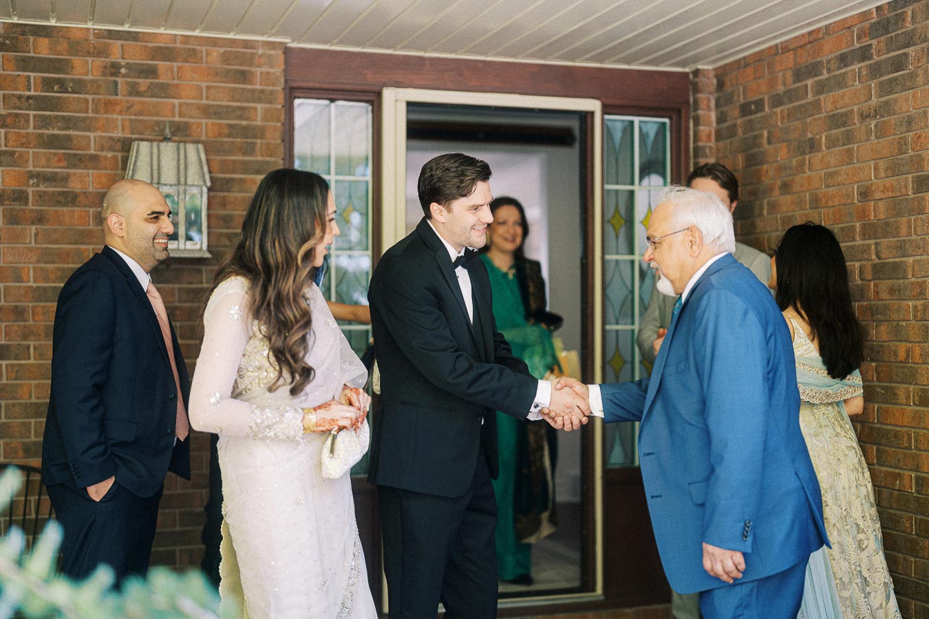 A group of people dressed in formal and traditional attire gathered at an event, with a man in a black tuxedo shaking hands with an older man in a blue suit in front of a brick wall and a door with stained glass panels.