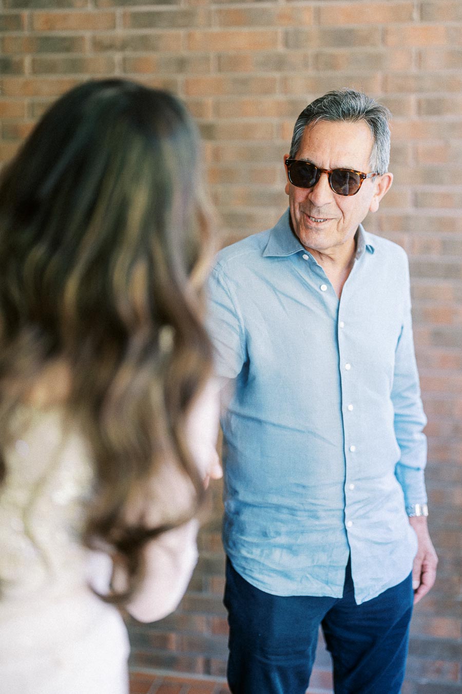 Man in a light blue shirt and sunglasses smiles while interacting with a person with long hair, standing against a brick wall background.
