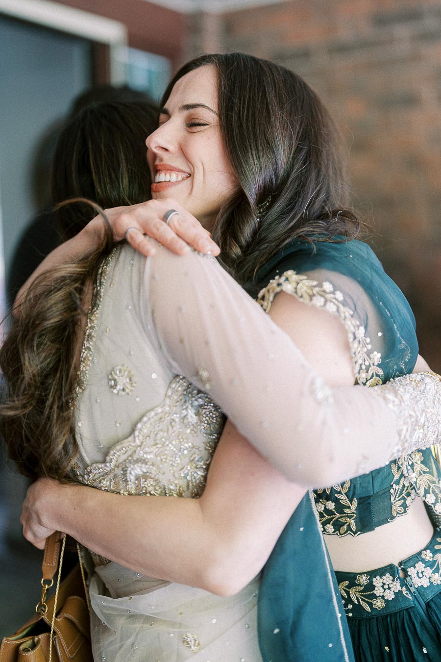 Two women happily embrace at a special event, wearing intricately embroidered outfits, reflecting joy and celebration.