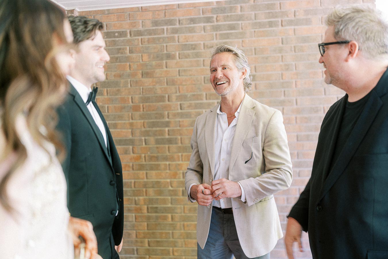 Group of well-dressed people having a conversation in front of a brick wall, focusing on a smiling man in a light suit jacket.