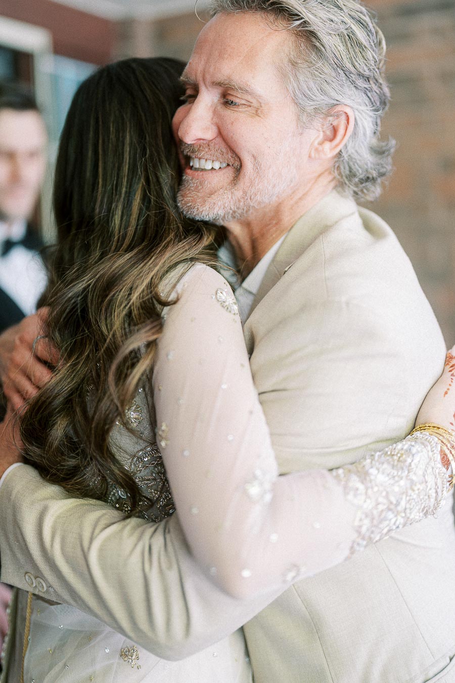 Smiling older man in a beige suit warmly hugging a woman with long brown hair and an embellished dress, capturing a joyful and heartfelt moment at a celebration.
