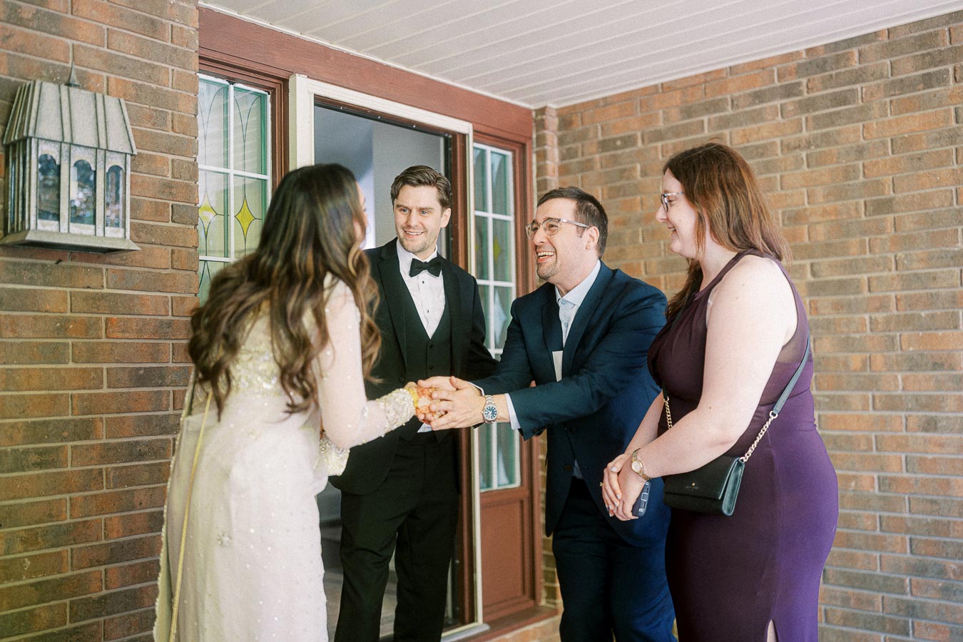 A group of four people, dressed in formal attire, warmly greeting each other at a front door with a brick exterior and decorative glass windows.