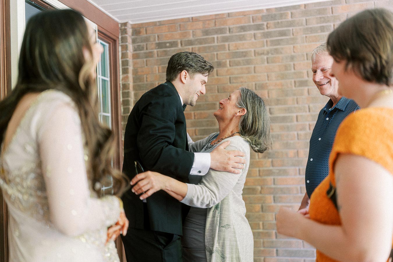 A joyful family moment with a group of people embracing and smiling warmly near a brick wall background, dressed in formal attire, capturing the essence of a celebratory gathering or special occasion.